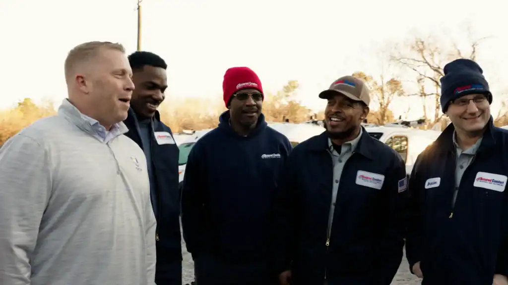 Five men wearing work uniforms stand together outdoors, smiling and talking. Vehicles and trees are visible in the background on a bright day, highlighting their camaraderie as they maintain close contact with one another.