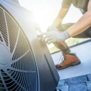 A technician wearing gloves and boots inspects or services an outdoor HVAC unit in bright sunlight, reflecting the dedication and professionalism highlighted in our About Us section.
